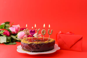 Selbstgebackener Kuchen mit Schriftzug und Kerzen steht neben Blumen und einem Briefumschlag vor rotem Hintergrund.